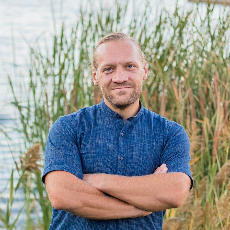 A man named Tim Vreeland standing in front of a lake with reeds, enjoying the serene beauty of nature.