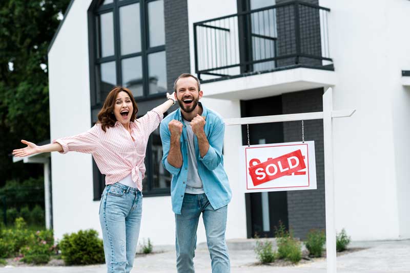 A couple stands proudly in front of their new home, featuring a sold sign, representing Murray's lighthouse home selling solution.