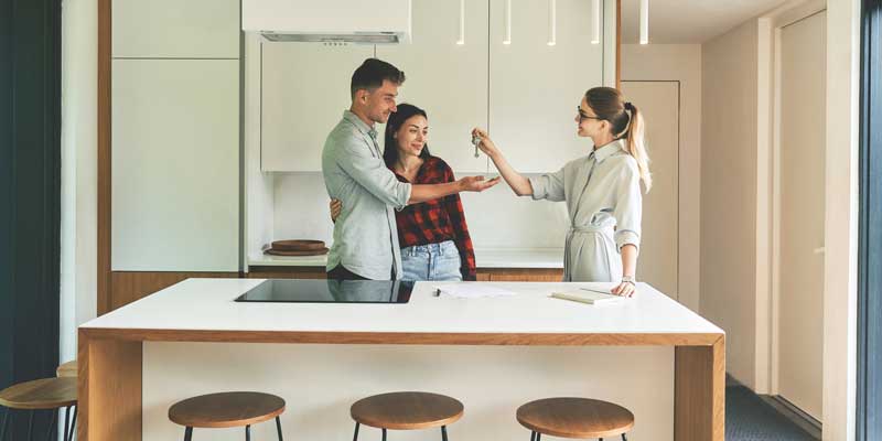 A couple stands in a kitchen with a white countertop, discussing home selling solutions in a lighthouse-themed setting.